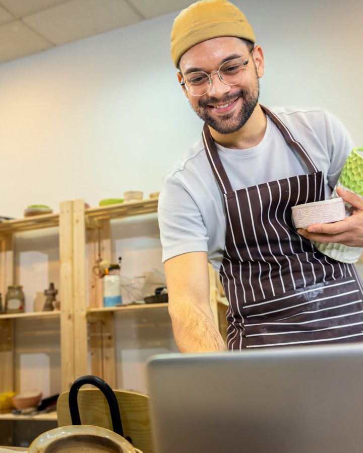 Male business owner using laptop in pottery studio workshop.